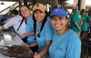 Three smiling students in an outdoor classroom with a large fish on the table