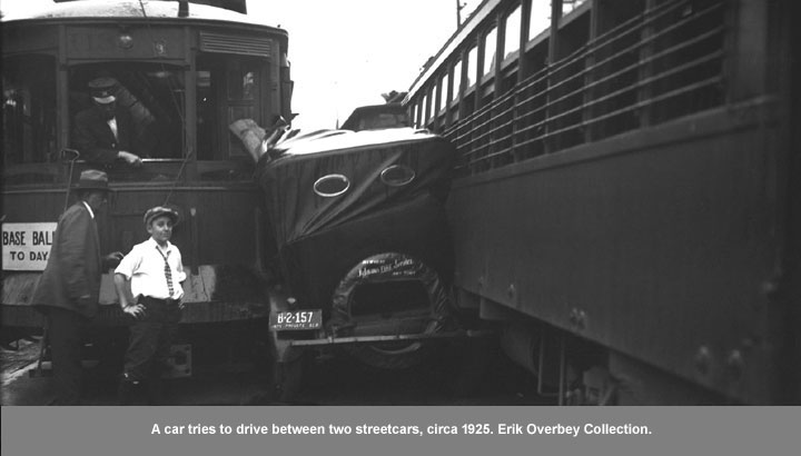 Black and white image of a car wedged between two streetcars.